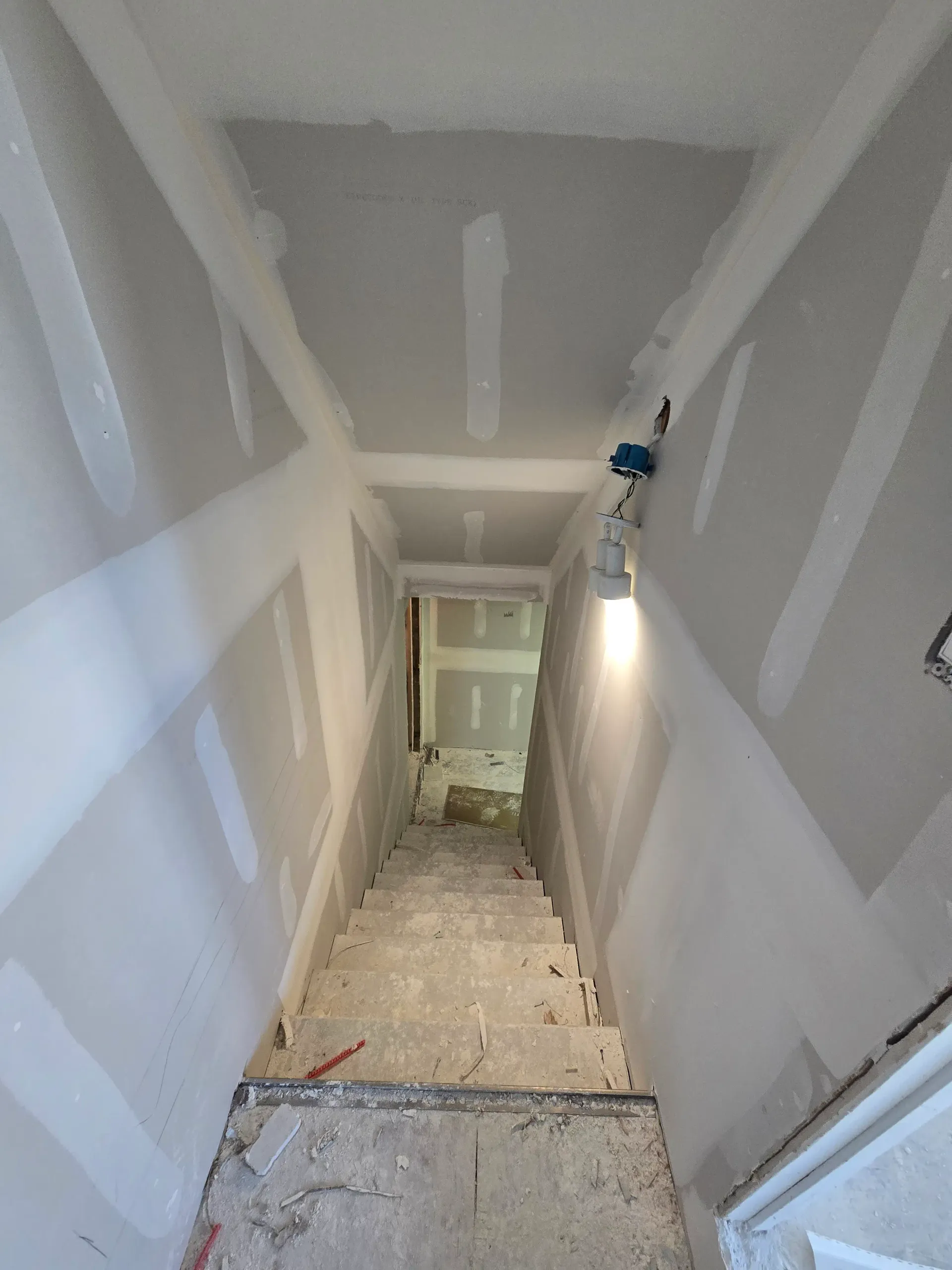 Staircase with newly installed and taped drywall under temporary lighting.